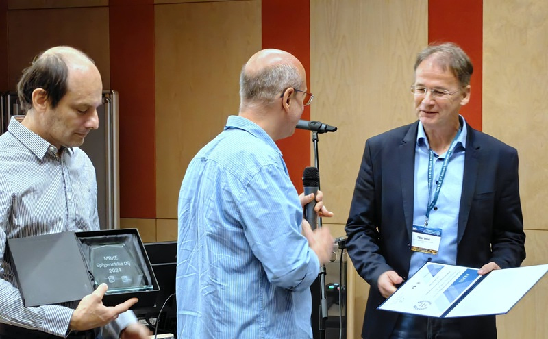 Three men standing, with the one on the right holding a certificate, the one in the middle a microphone, and the one on the left a trophy.