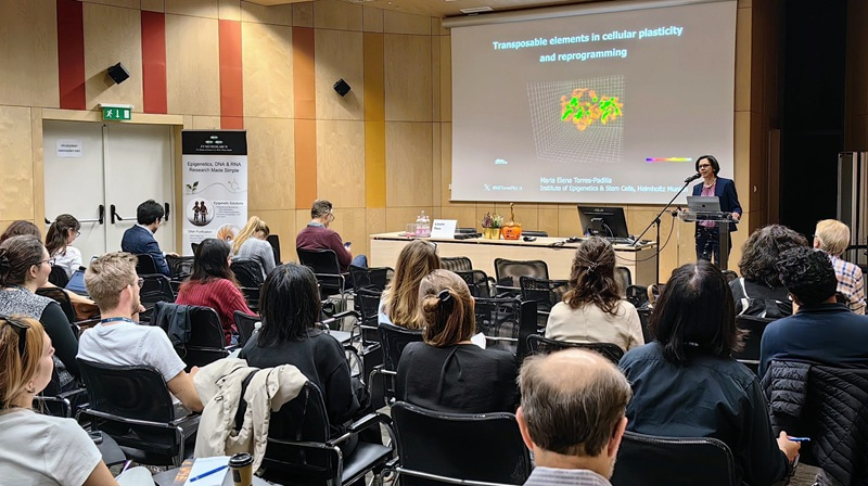 Photo from the audience of the speaker standing at the front of the room presenting her slides.