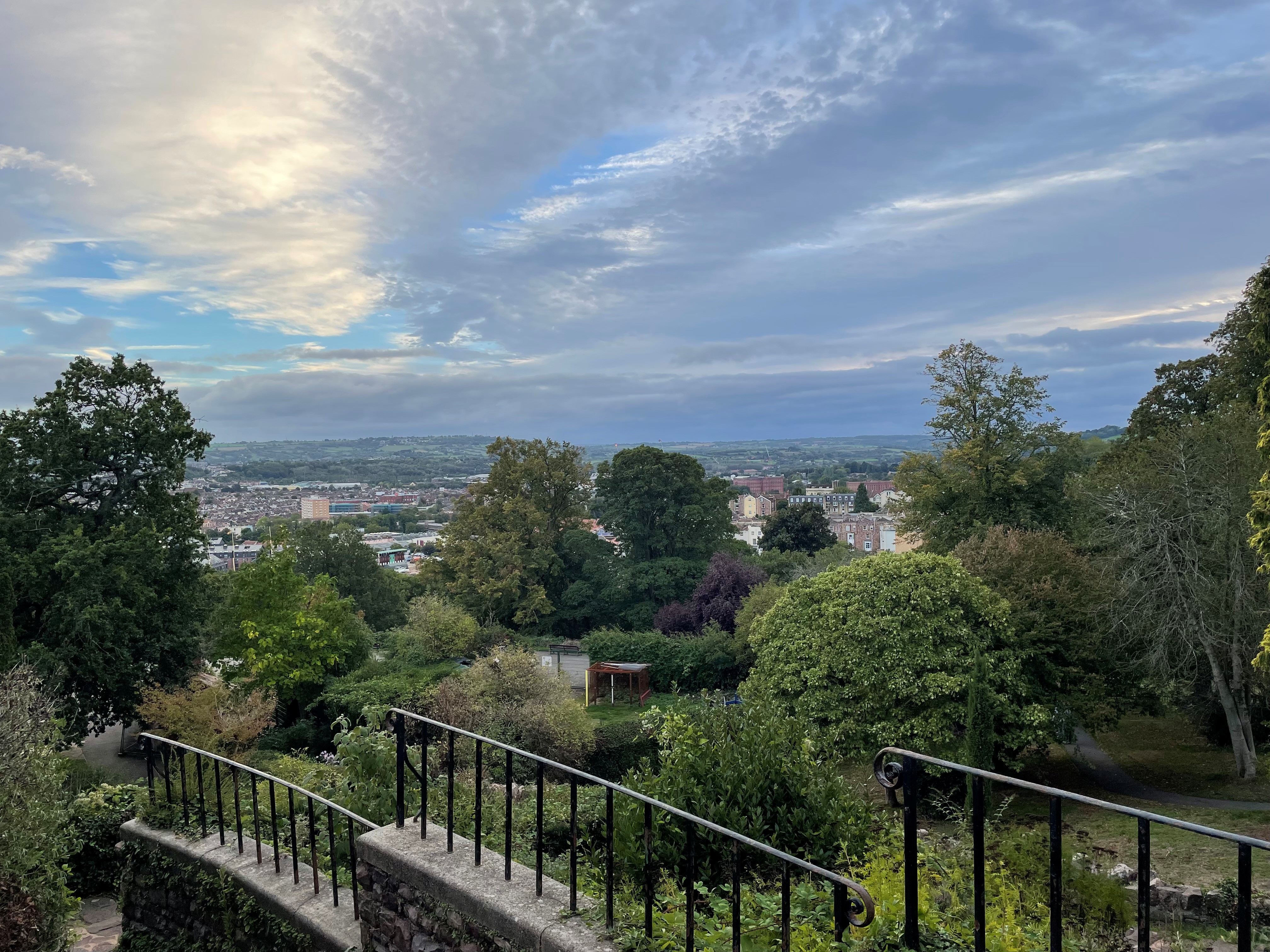 View from the base of Cabot Tower in Brandon Hill Park, Bristol. Steps descend through trees and gardens towards the city, with Bristol's skyline and surrounding hills visible under a cloudy sky.