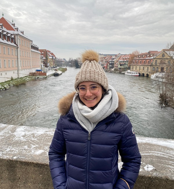 Photo of Sara León Álvarez, smiling to the camera and standing on a bridge over a river in Bamberg, Germany.