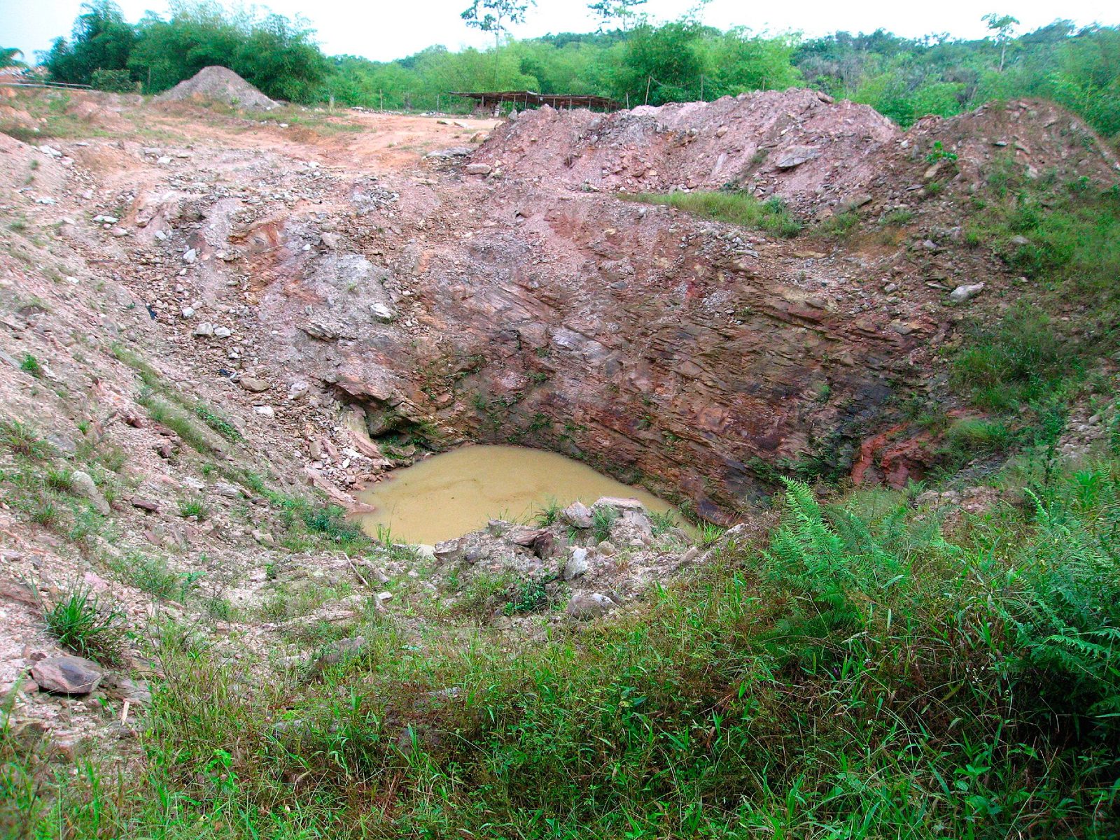 Bare, reddish mining pit holding standing water, surrounded by disturbed soil and regrowing grass.