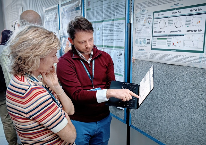 A man holding a laptop and pointing at the screen, with a woman standing next to him and looking at the laptop.