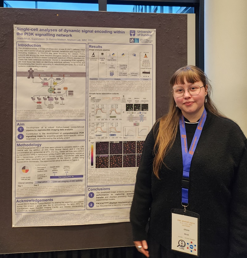 Oliwia Mruk wearing a black jumper and a lanyard, standing in front of her research poster.