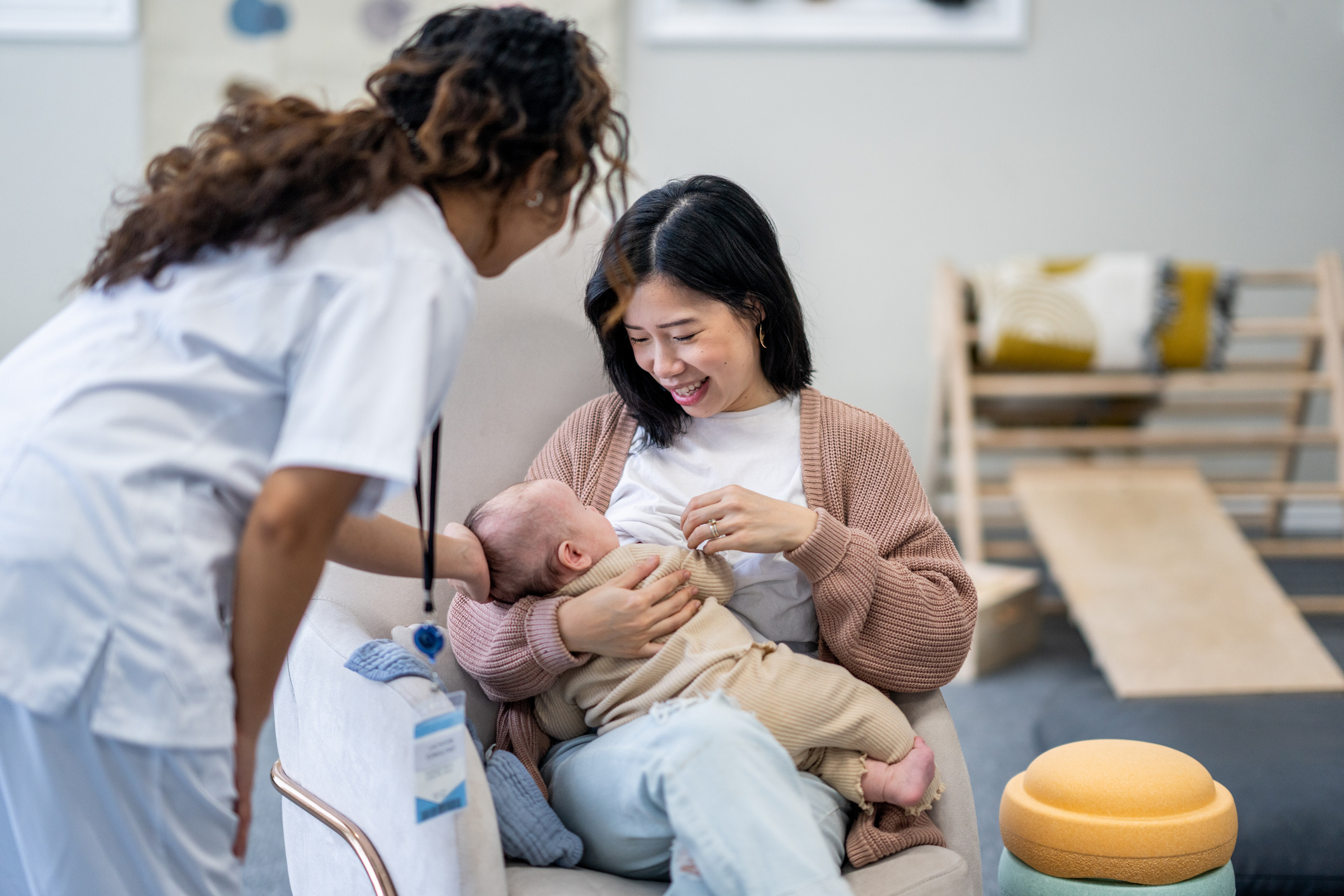 An asian woman sitting in a chair and breastfeeding a baby. A dask skinned lactation consultant supporting her and discussing the breastfeeding process