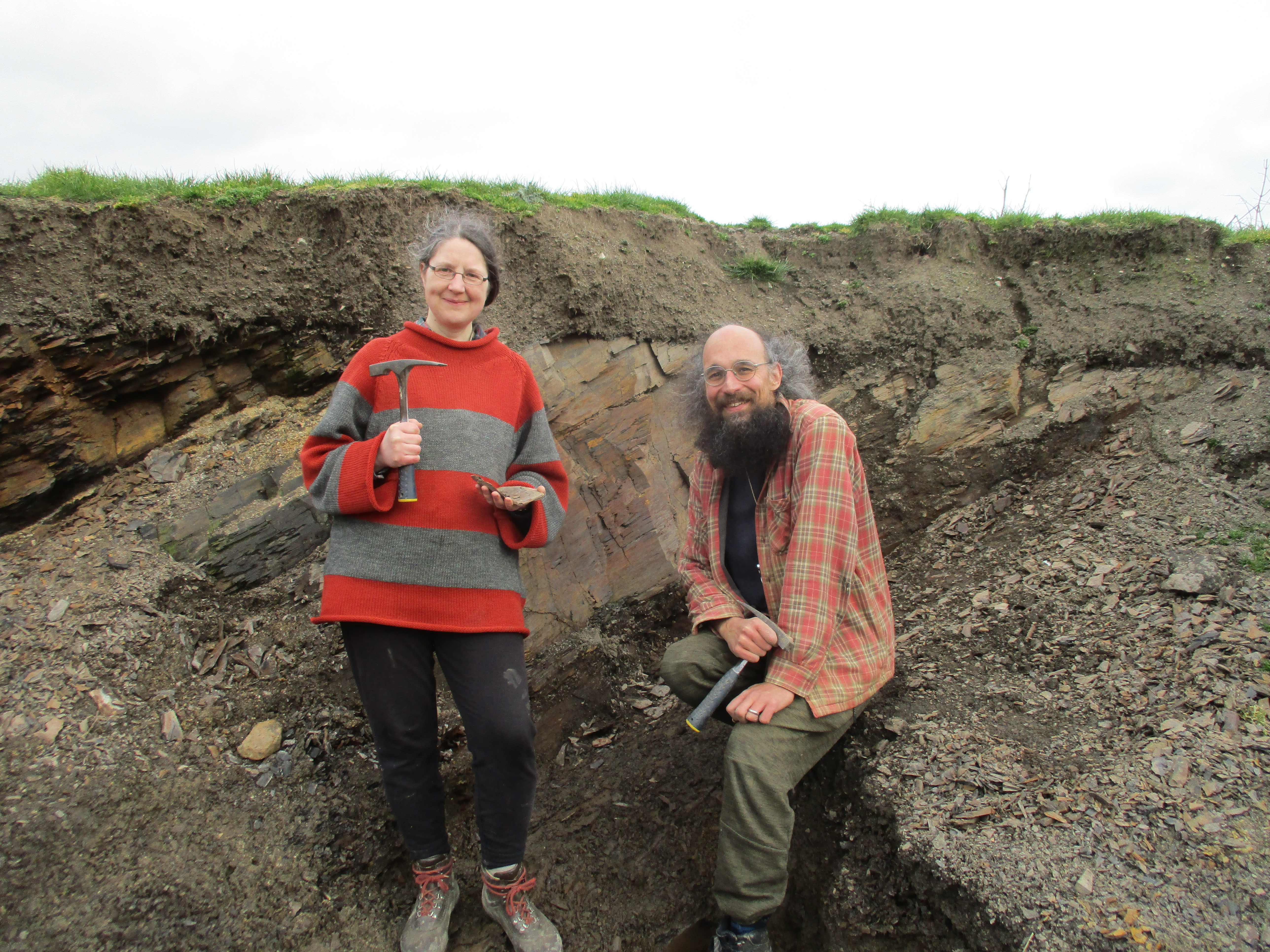Two happy palaeontologists (the authors of the article) holding their hammers in the quarry. It's a very small rock face with the beds (including a white volcanic ash layer) dipping down to the left. 