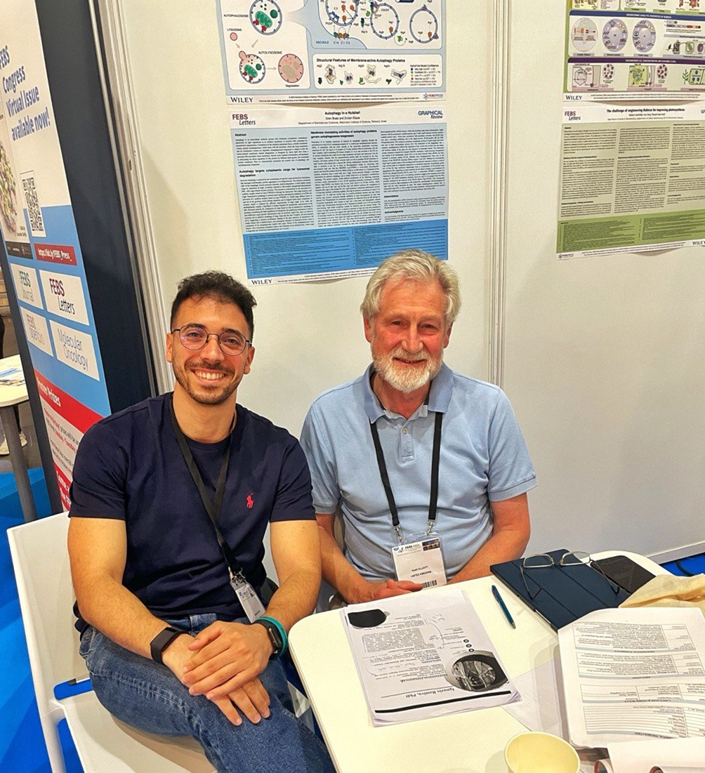 Photo of Ignazio Restivo and Dr. Keith Elliot, sitting down and smiling at the camera, with scientific posters on the wall behind them.