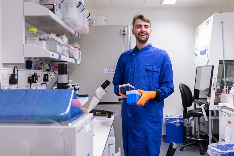 Student in a lab facing the camera, holding plastic boxes, and smiling.