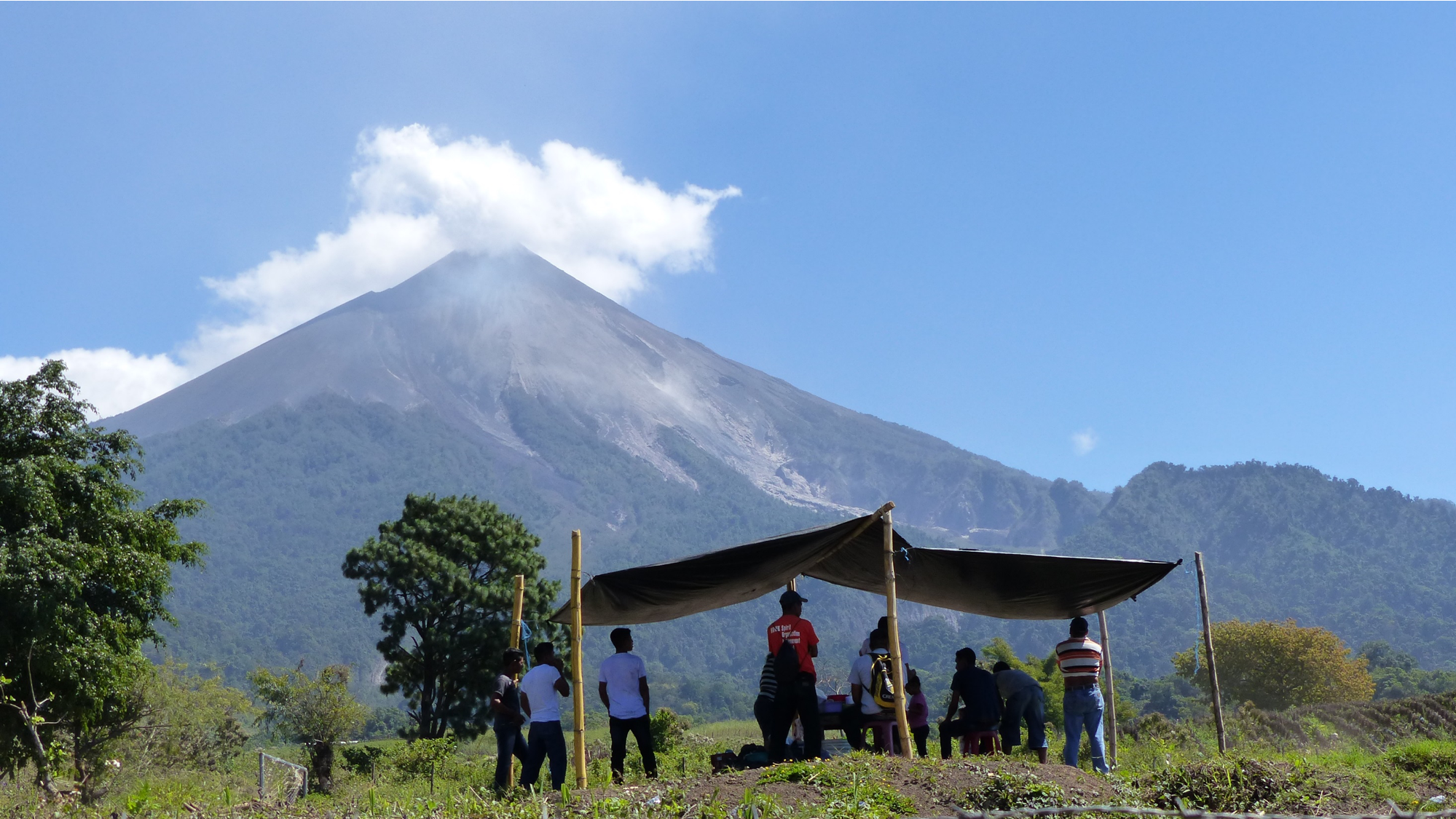 A photograph of people standing under a shelter in front of Fuego volcano, Guatemala.