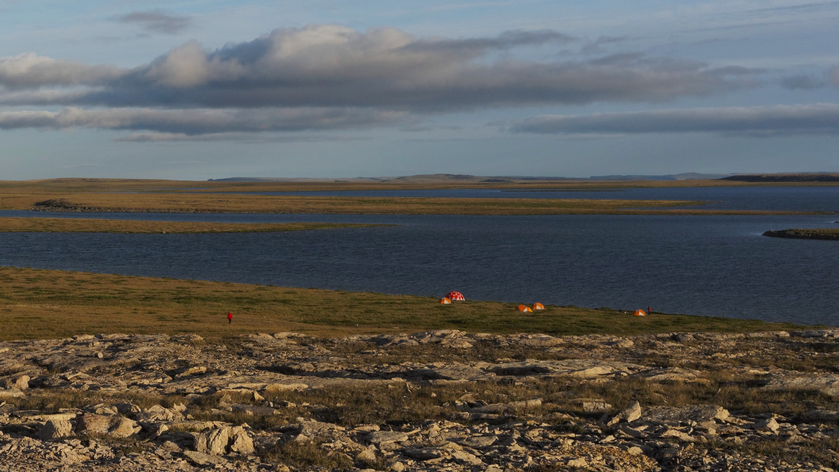 A lake situated within a vast, treeless Arctic landscape. Researchers are camped out in tents on the shore, and one of them is seen standing in the middle of the landscape.
