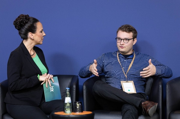 Photo of Johann Liebeton talking to a woman, both of them sitting down on sofa chairs in an interview setting.