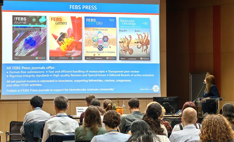 Photo from the audience of the screen and the presenter on the right looking at the slide.