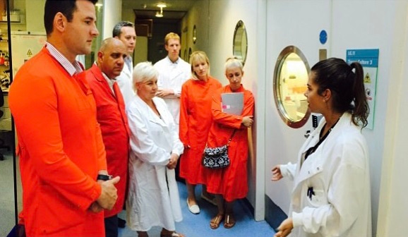 Photo of a group of seven visitors to a laboratory, all wearing lab coats, listening to Charlotte Bellamy, who is facing them.