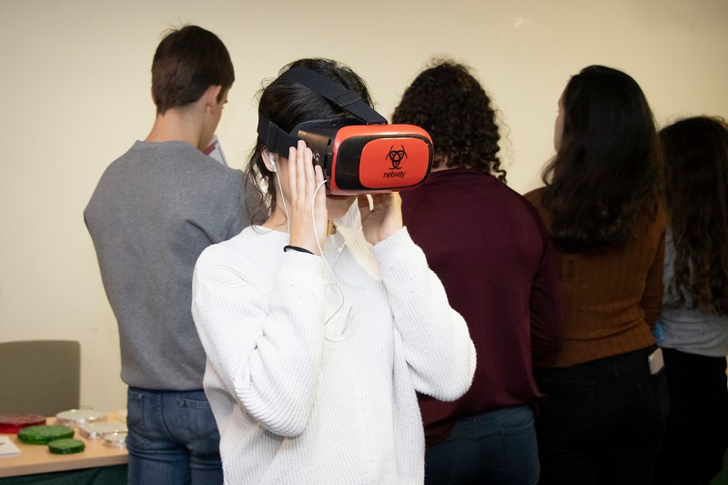 Girl looking into a Virtual Reality headset, facing the camera, with other young people behind her.