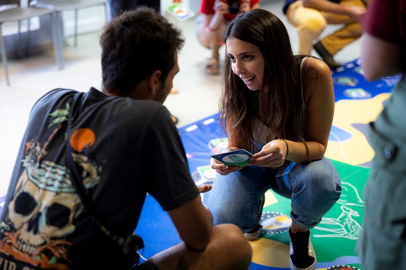 Photo of two people kneeling down and talking to each other. One is a young woman facing the camera, holding a card and talking to a youg man who has his back to the camera.
