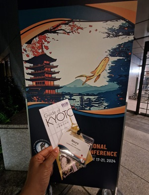 Photo of Noura's hand holding the conference pass and a leaflet in front of the event banner.