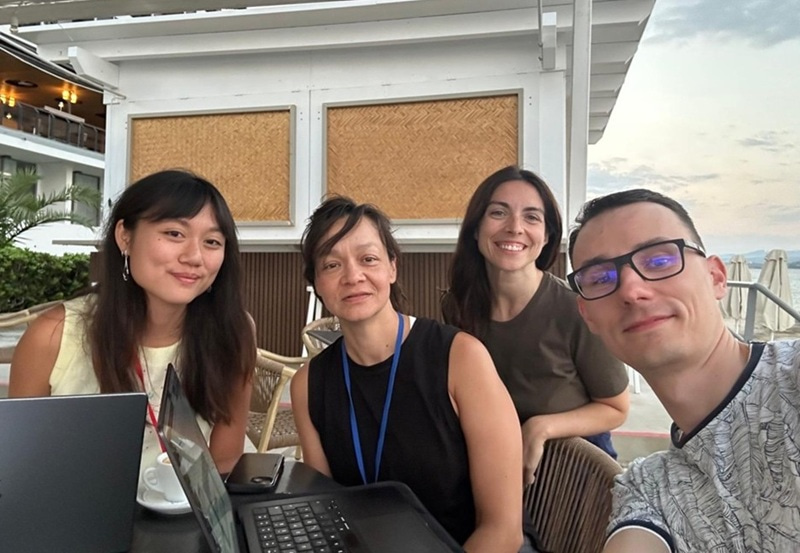 Four people sitting around a laptop and taking a group selfie.