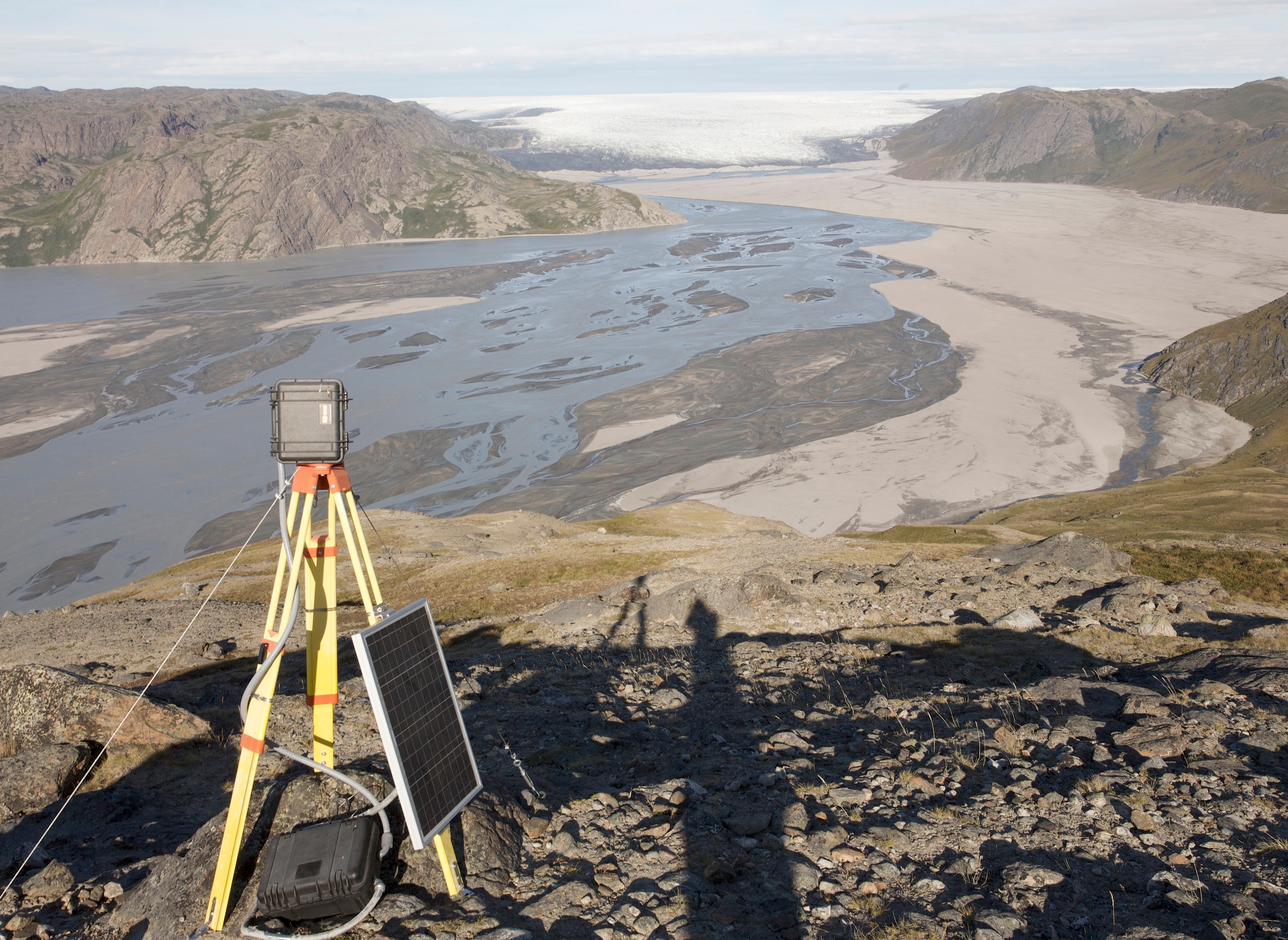 Photograph of a river emerging from the ice sheet margin flowing over tundra toward the ocean in southwest Greenland, with a remote time lapse camera monitoring station in the foreground.