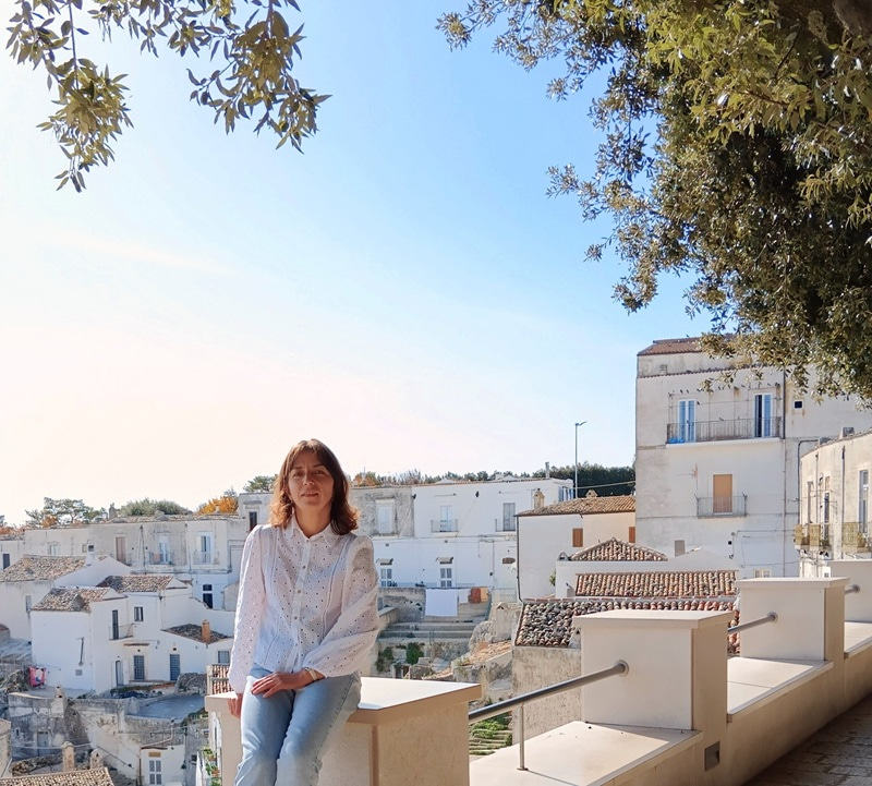 Photo of woman sitting and looking at the camera, with houses in the background and blue sky and tree branches over them.