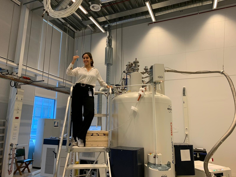 Photo of Cristina V. López Gallego standing on the stairs next to an NMR spectrometer.
