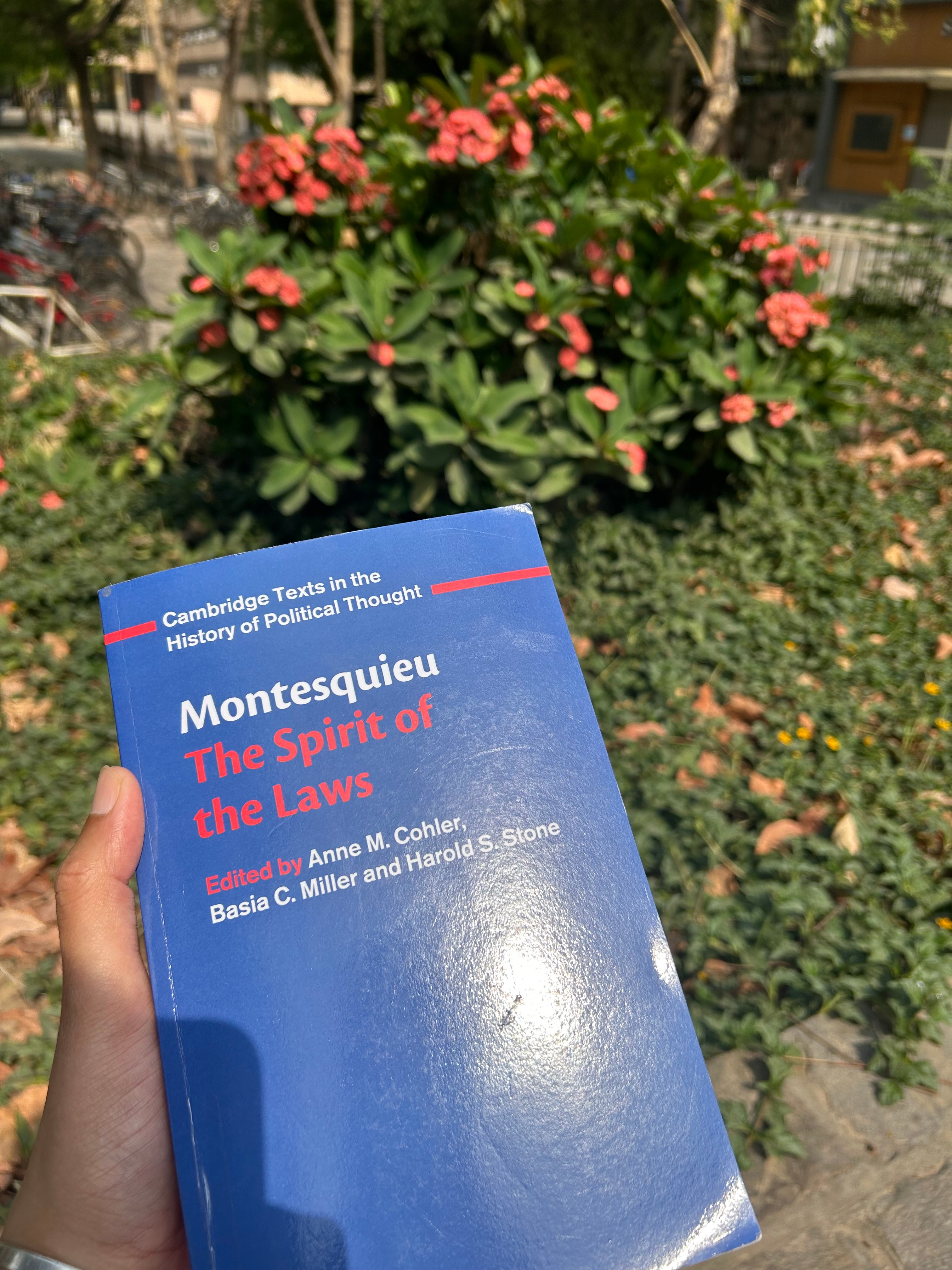 A hand holding Montesquieu’s "The Spirit of the Laws" in front of blooming coral-pink flowers on a green campus lawn.