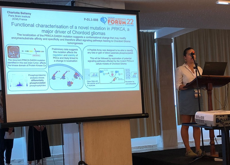Photo taken from the audience of Charlotte Bellamy's presentation at the Young Scientists Forum. The photo shows her standing behind a lectern and a screen to her right showing a slide about her work.