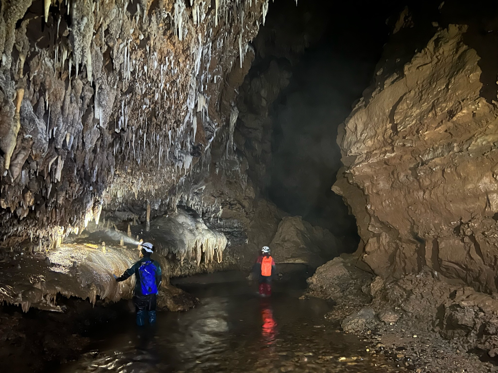 Co-authors Don Rodbell and Liz Olson walking along the river in Huagapo Cave. To the left of the image is a bench with multiple speloethems. 