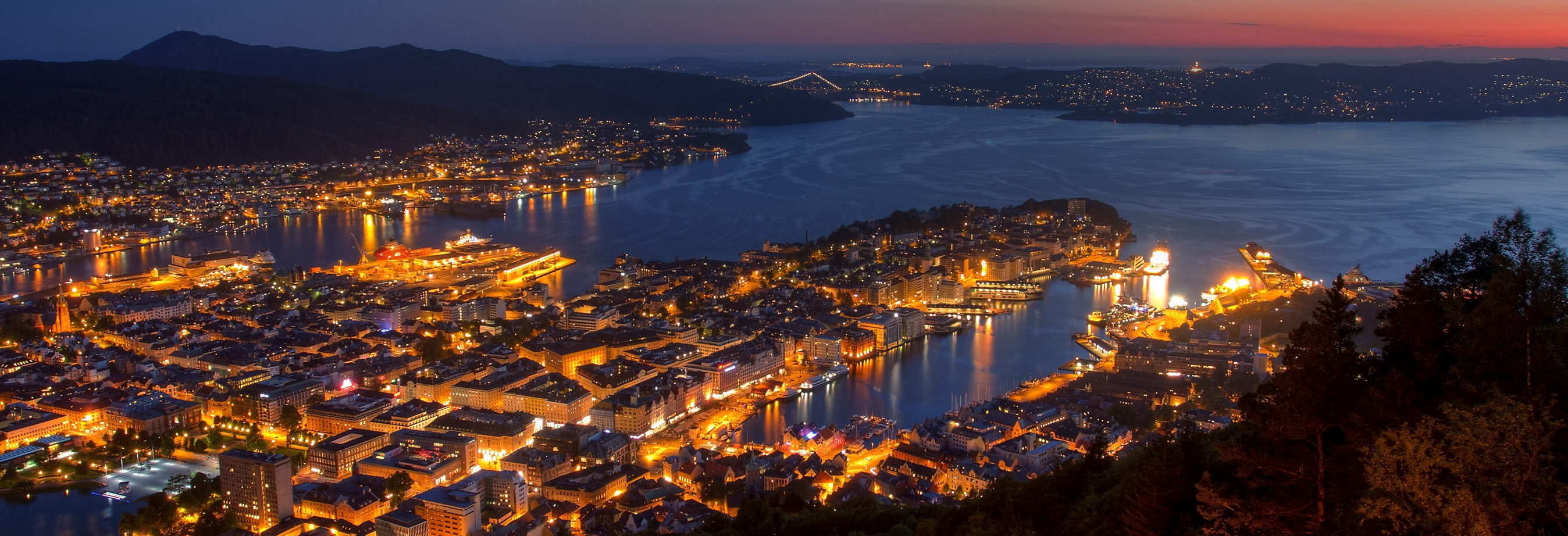 Night view of Bergen, Norway, from Mount Fløyen, showing the illuminated city, harbour, surrounding fjords, and mountains at dusk.