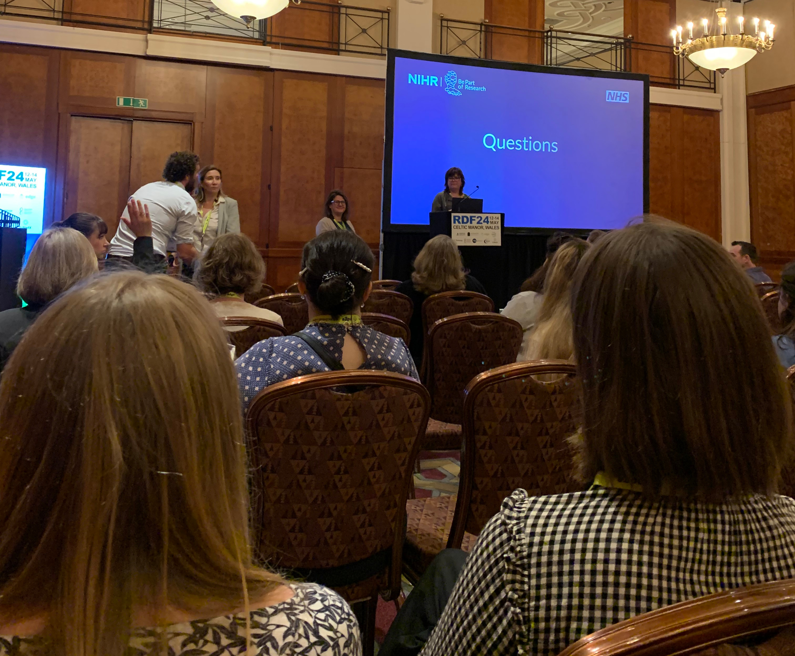 audience looking at a screen with the word 'Questions' and a person with a hand raised