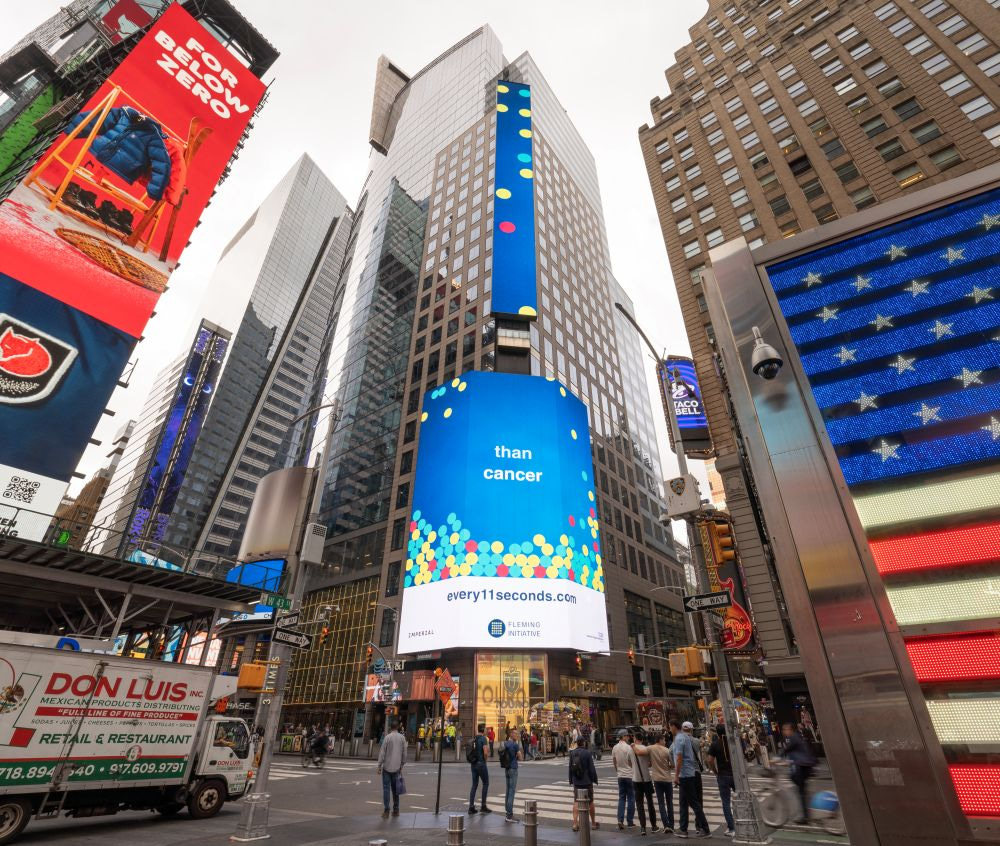 A busy street scene in Times Square, New York City, featuring towering digital billboards. The central billboard displays a text excerpt from an animated public health message on a blue background with colorful dots. At the bottom of the billboard on a white background text reads “every11seconds.com”.