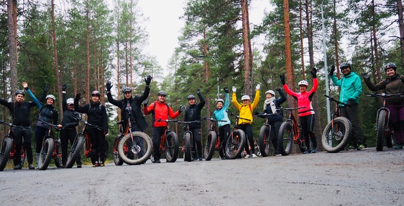 Large group standing over their bikes on a forest road.