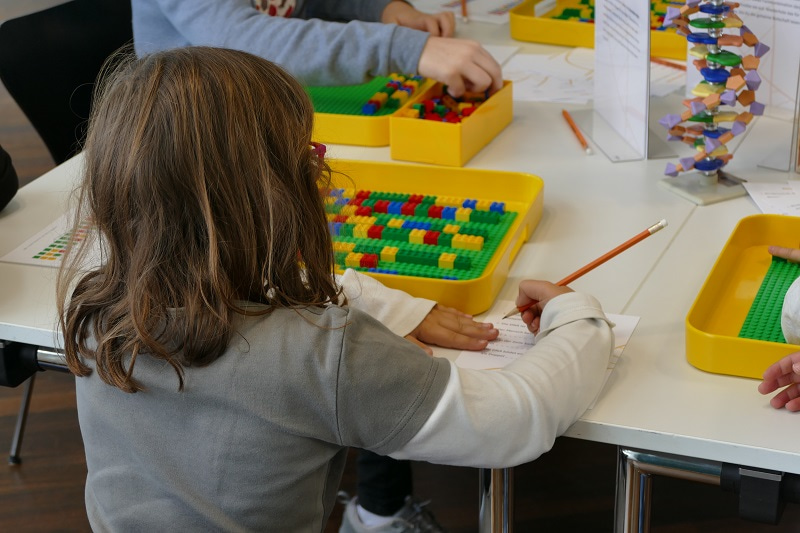 Photo of a young girl with her back to the camera and sitting at a table, writing on a paper.