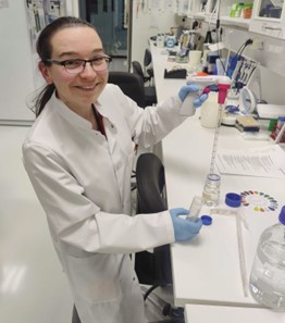 Photo of Anja Hartewig in the lab, smiling to the camera and holding a pipette.