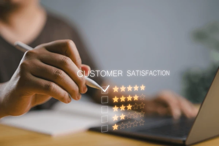 Hand with a stylus checks a five-star "Customer Satisfaction" rating on a screen near a laptop. Focus on digital feedback, tech setting.