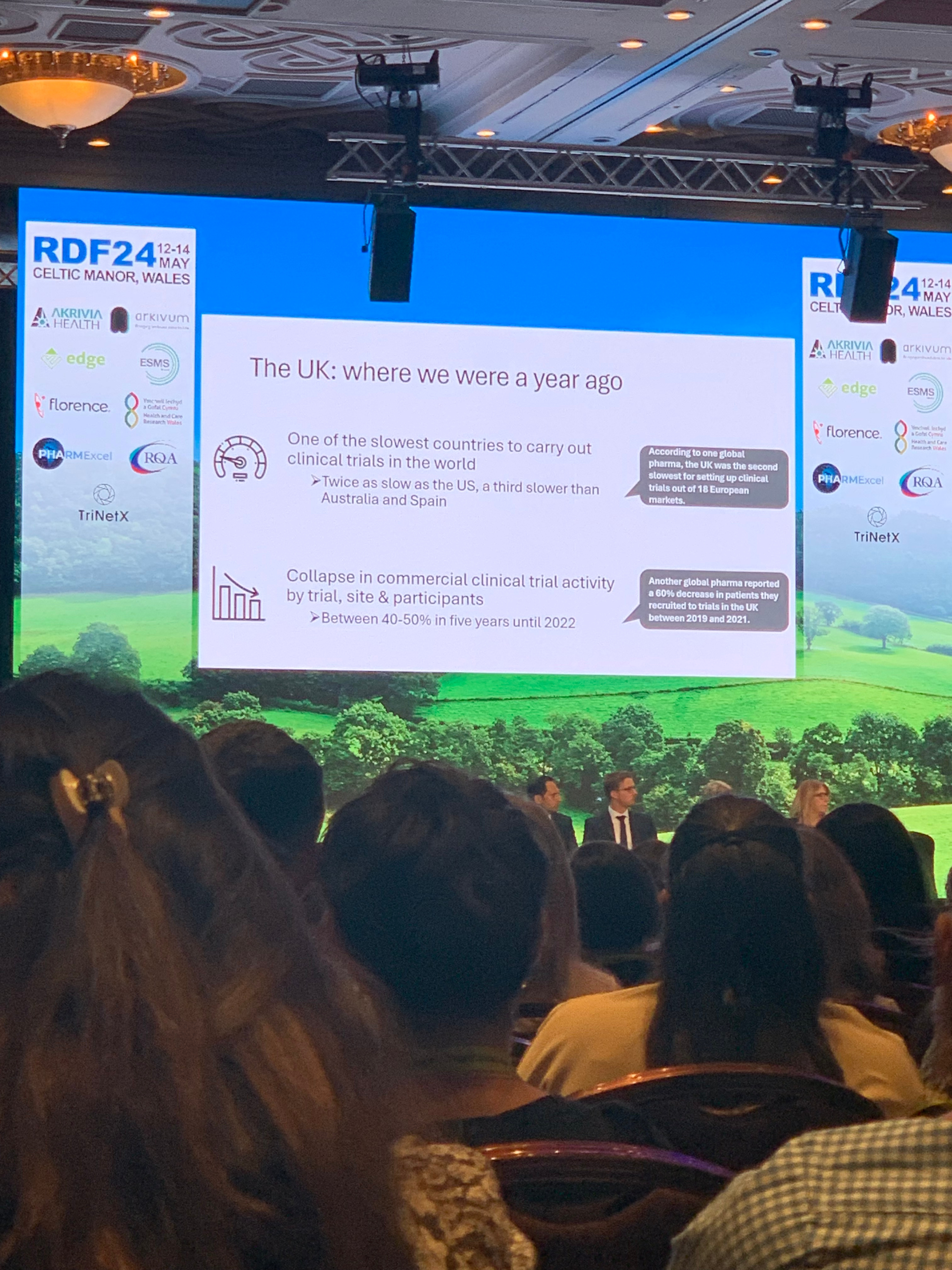 audience looking at people on a stage with a presentation on a big screen with the caption 'The UK: where we were a year ago'