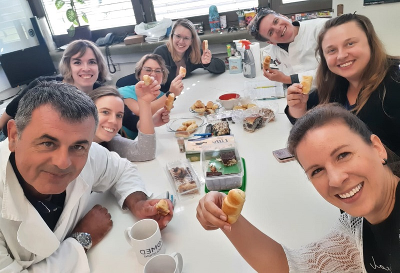 Photo of a group sitting at a table, and looking up at the camera while holding food and smiling.