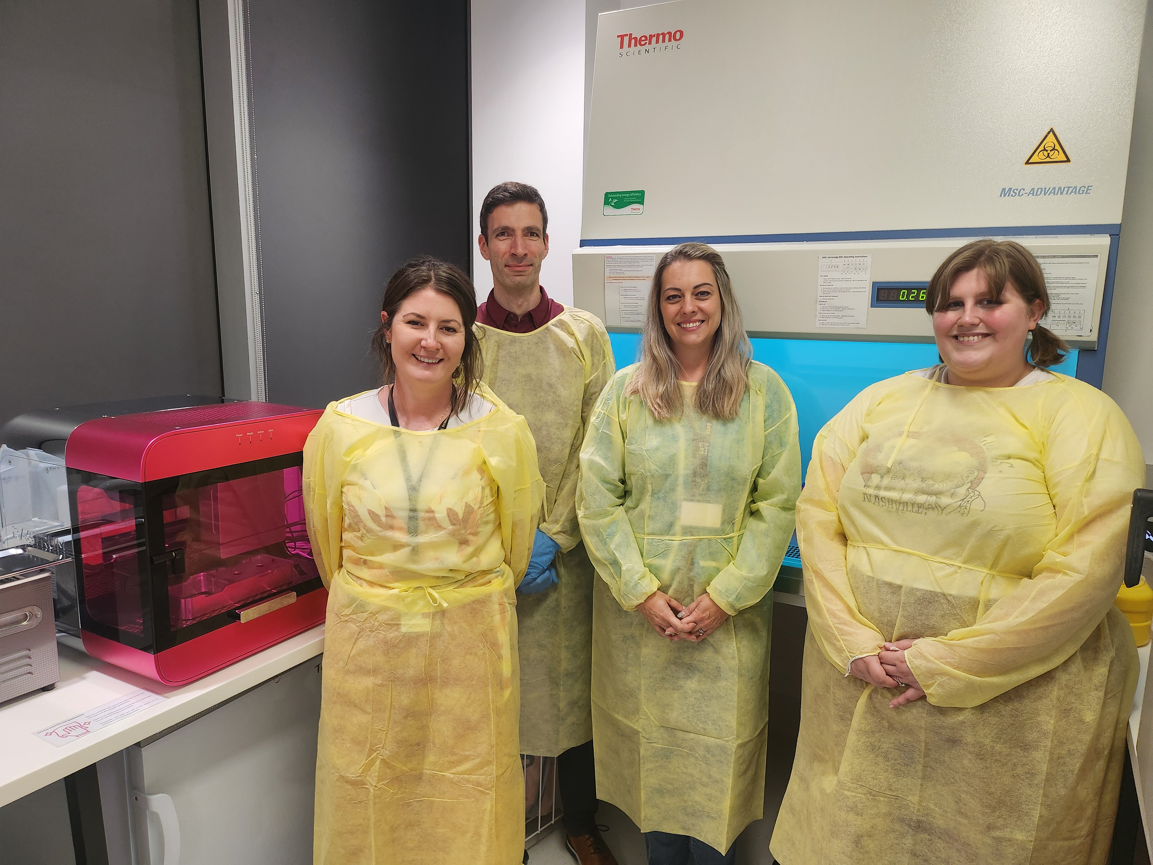 Researchers stand in yellow lab coats next to a pink RASTRUM bioprinter. Order of researchers from left to right: Dr Claire Richards, Dr Martin Engel, A/Prof Lana McClements and Miss Ashley Bannister.