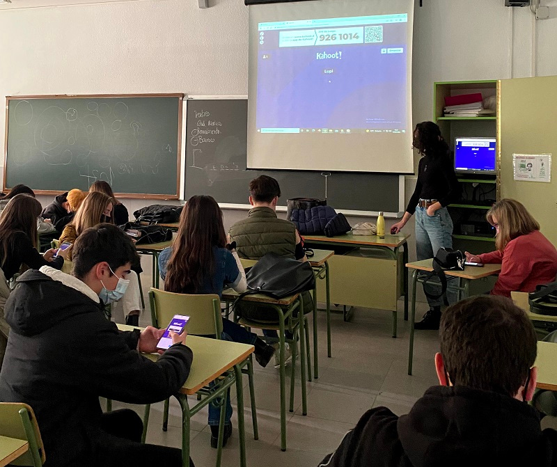 Photo taken from the back of a classroom, showing Clara Rey presenting an online survey to young people, who are checking the results on their phone.