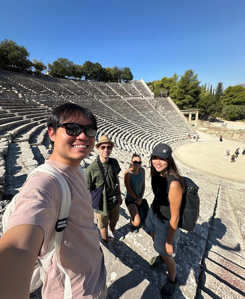 Four young people taking a group selfie with the steps of an amphitheater below.