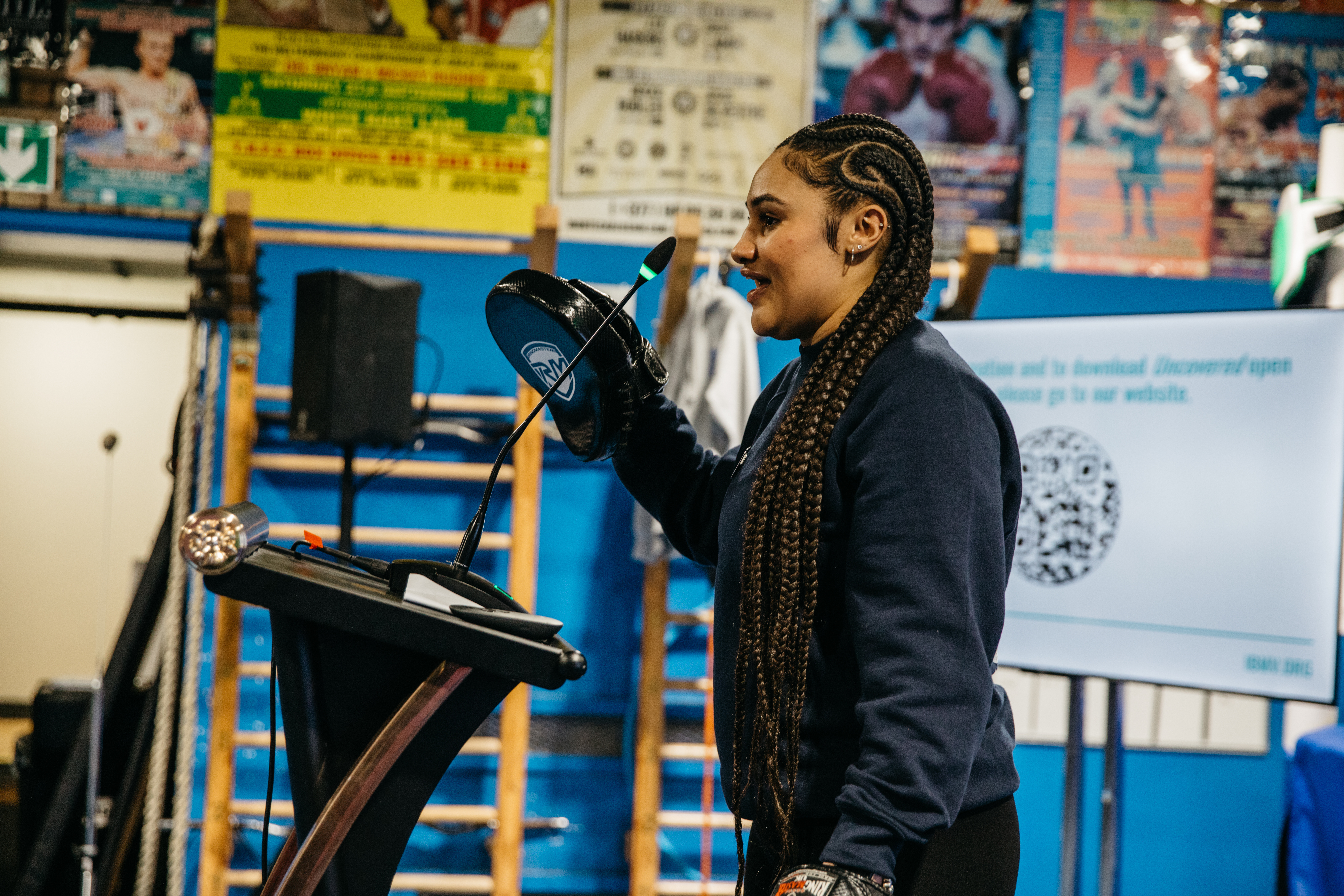 Woman holds boxing gear up while speaking at a microphone.