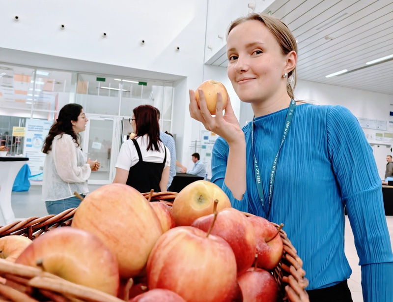 Photo of a young woman holding an apple and smiling at the camera, with more apples on the foreground.