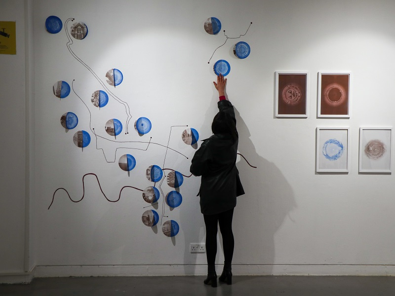 Photograph of a person from behind reaching up to touch artwork displayed on a wall. Created by Carla Fernández Arce in collaboration with Richard Colechester, Zhe Lou, Semyon Bodian, Fraser Watt and Shaoyan Zhang, a group of people with experience of heart scans, the Wellcome / EPSRC Centre for Interventional and Surgical Sciences at University College London and the University of the Arts, London Post Graduate Community. Creative commons licence CC BY-SA 4.0.  Photograph taken by Catriona Mahmoud, University of the Arts, London.