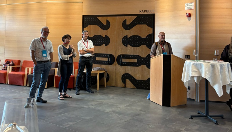 Photo showing four people standing next to each other and smiling at the audience, which is off camera.