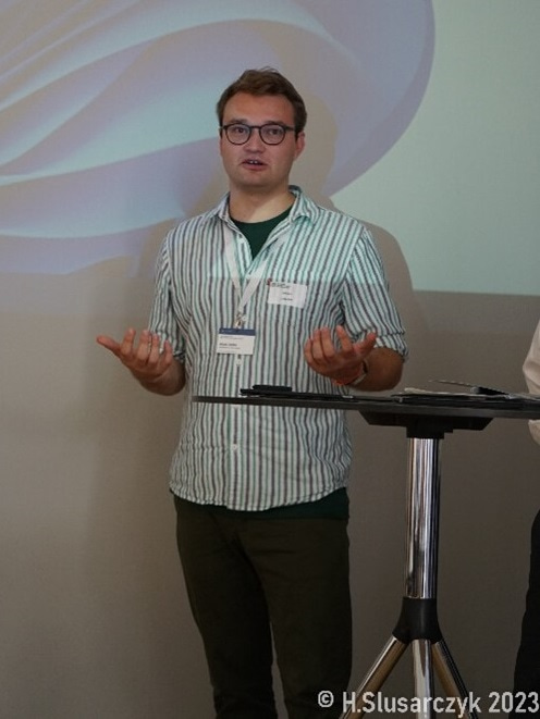 Photo of Johann Liebeton speaking at an event, standing by a tall chair and holding his hands out.