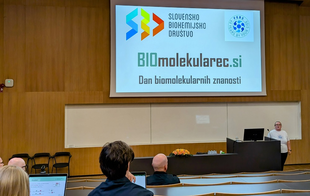 Photo of a young woman presenting on the stage, by a slide of the event.