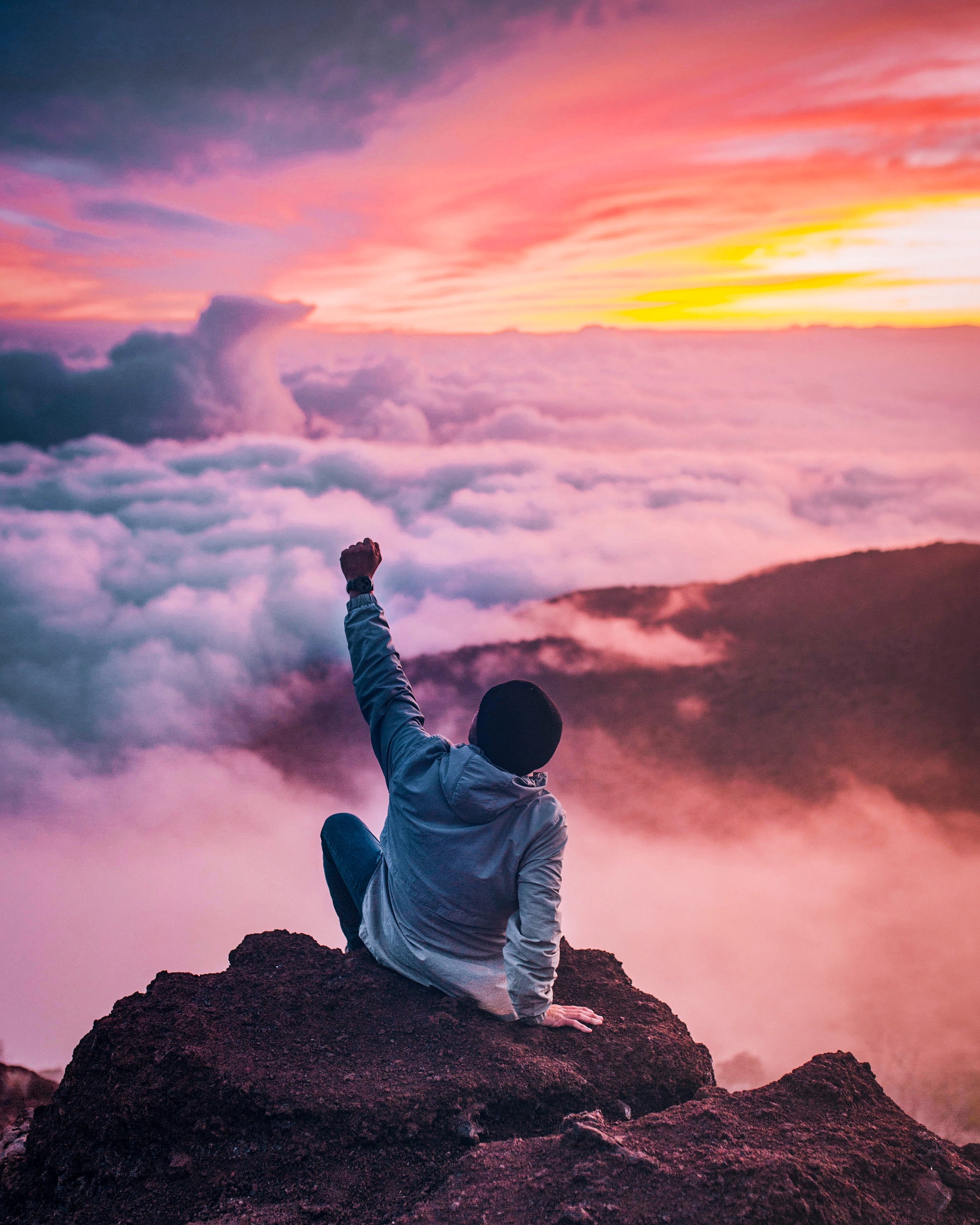 A man is sat on a mountain cliff, cheering with one arm. Soft clouds are below him and a beautiful sunset is before him. Photo by Ian Stauffer on Unsplash.