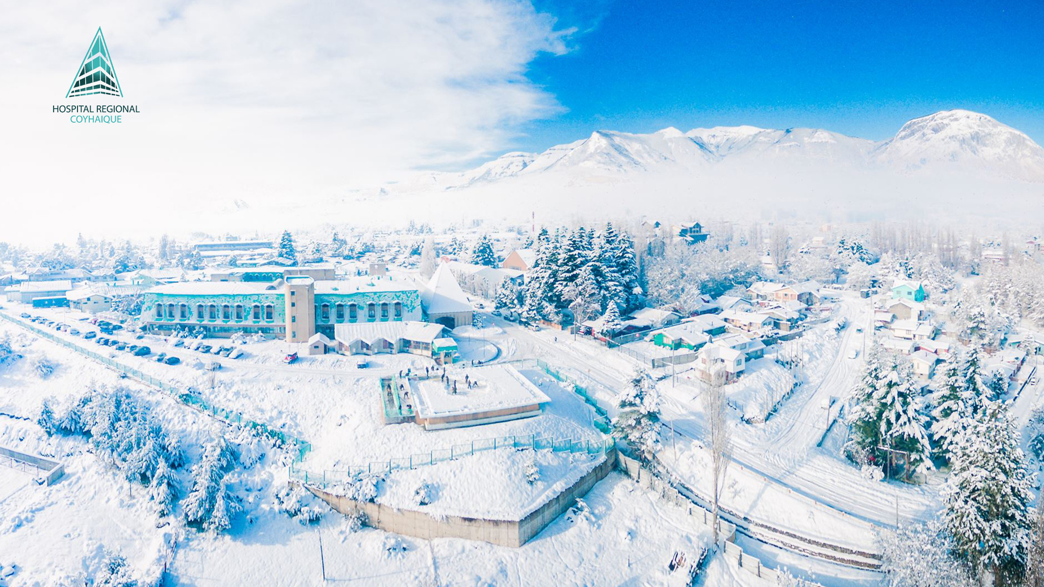 Aerial view of the Regional Hospital of Coyhaique covered in deep snow, surrounded by houses, trees, and mountains under a bright blue sky. The image shows the isolation and harsh winter conditions of southern Chile.