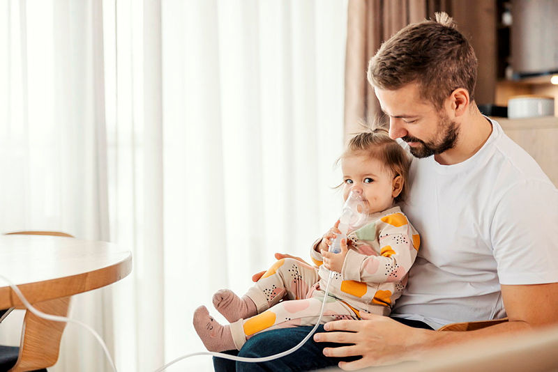 A child is inhaling medicine using a nebulizer sat on her father's knee.