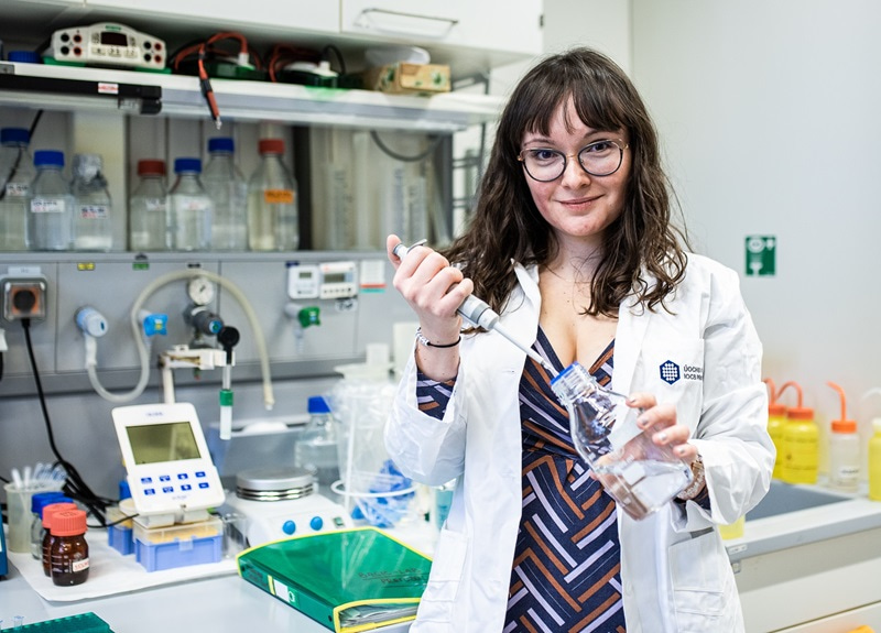 Photo of Veronika Vetýšková in the lab, holding a bottle and a pipette and smiling at the camera.