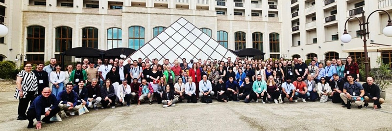 Photo of large group, with a glass pyramid structure and a building on the background.
