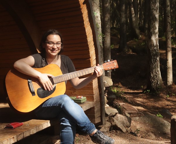 Photo of Anja Hartewig playing a guitar and smiling at the camera, sitting at the front of a cabin in the forest.